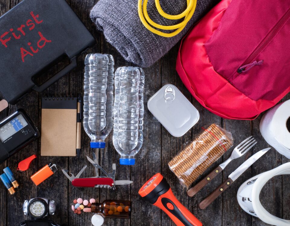 An assortment of survival gear necessary for natural disaster survival including water bottles, backpack, flares, and more. Bright colored items on a black background.
